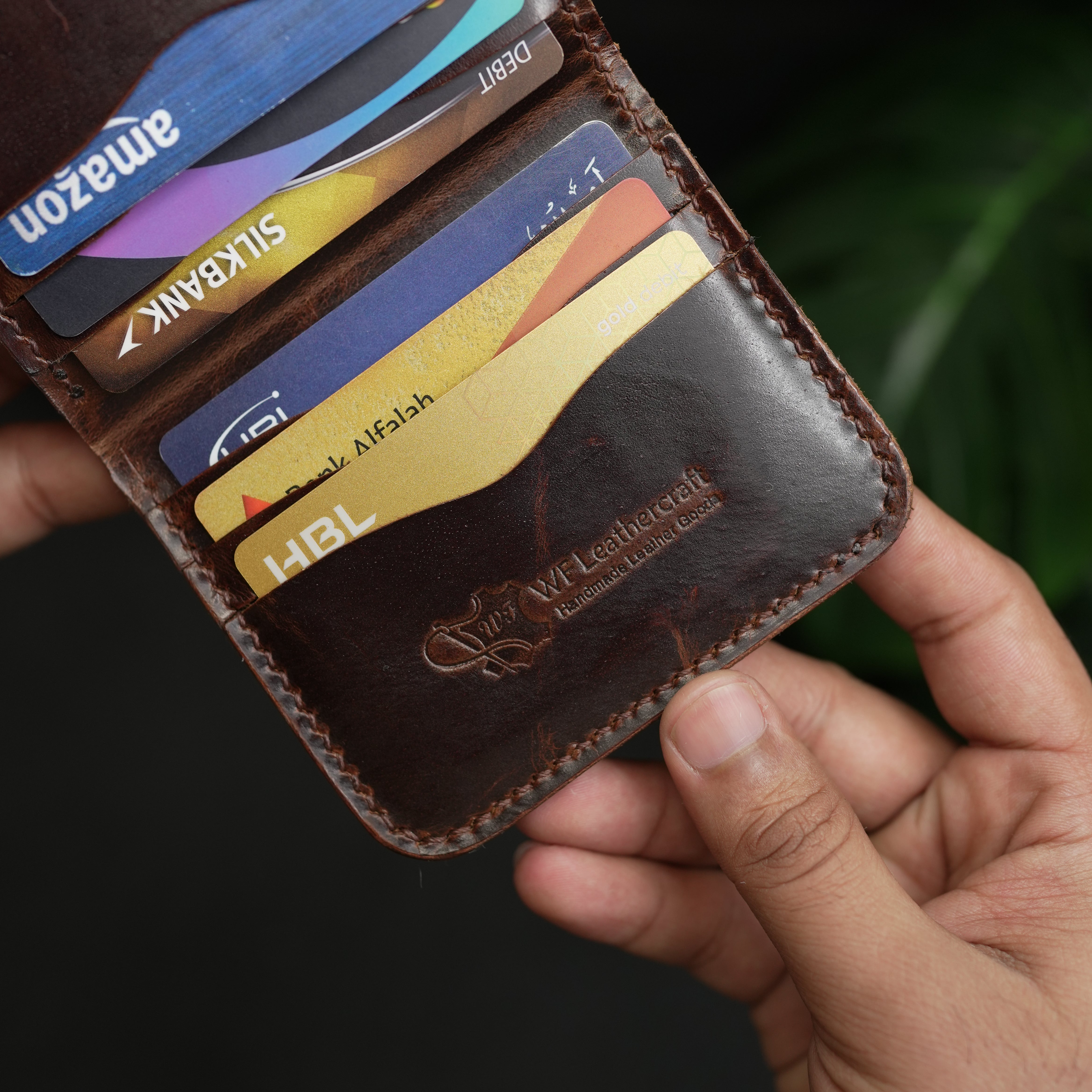 Hand holding a leather wallet with multiple cards, including Amazon and Silkbank cards.
