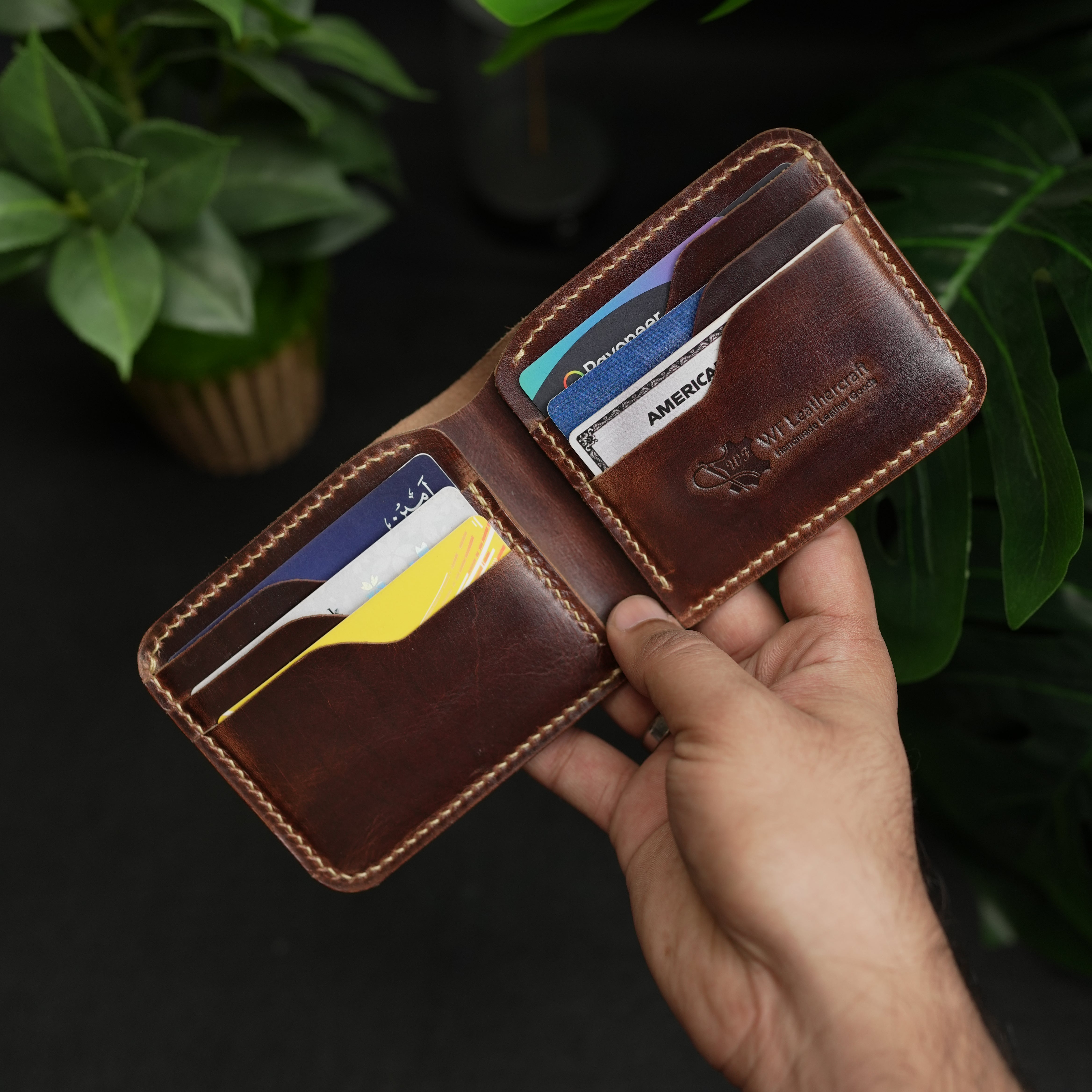 Brown leather wallet with card slots held by a hand against a dark background with green leaves.