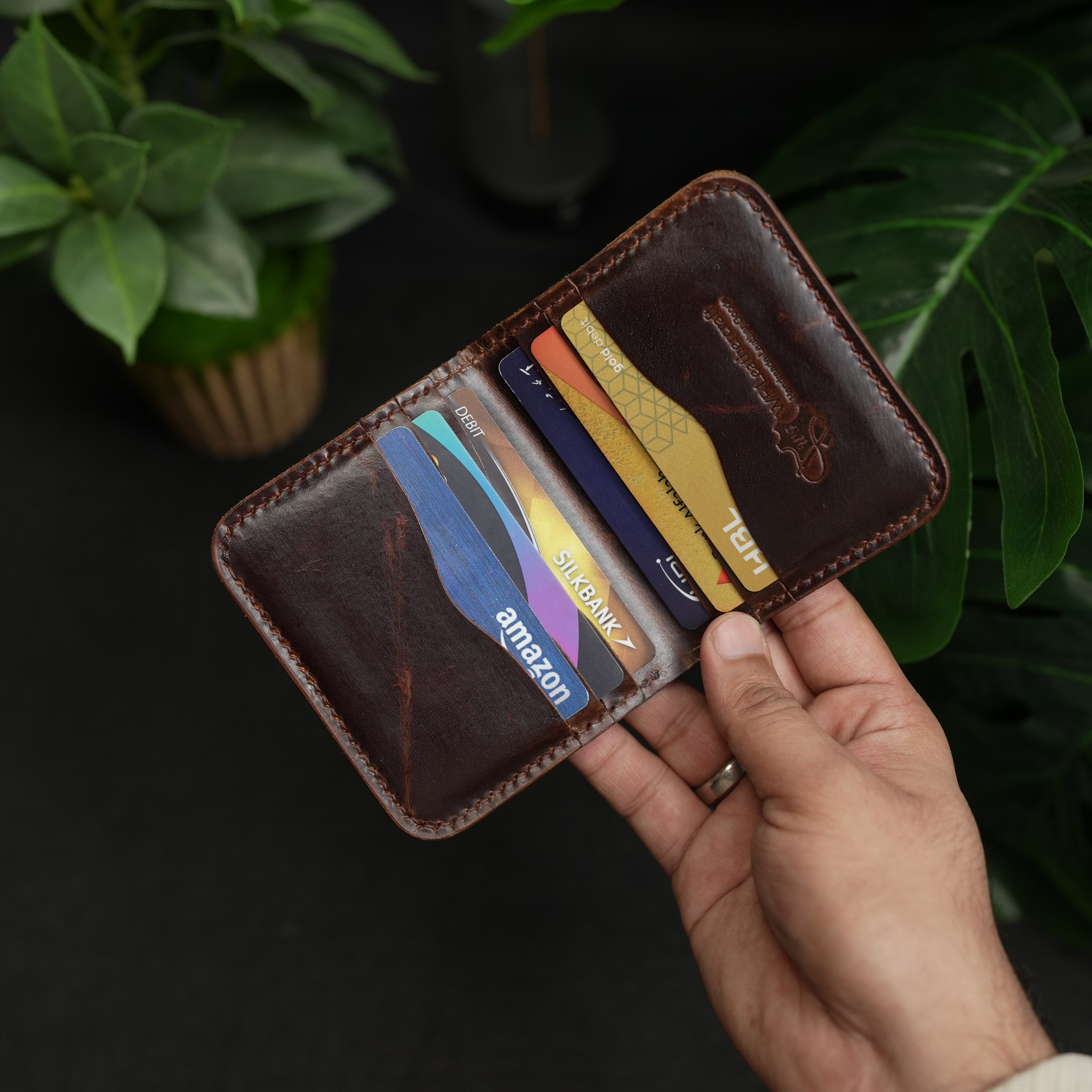 Hand holding a leather wallet with multiple cards against a dark background with green plants.