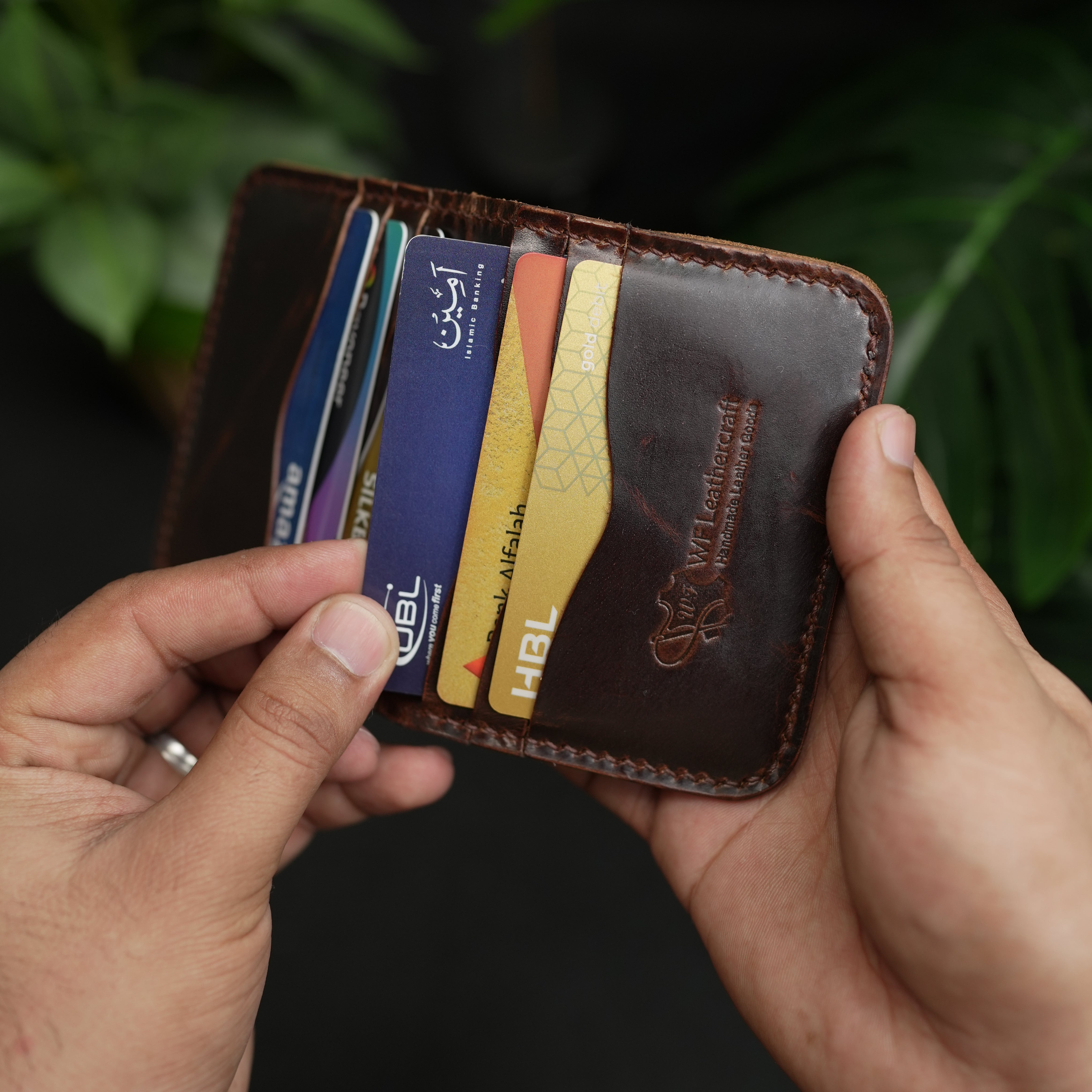 Hand holding a brown leather wallet with multiple cards inside, against a blurred green background.