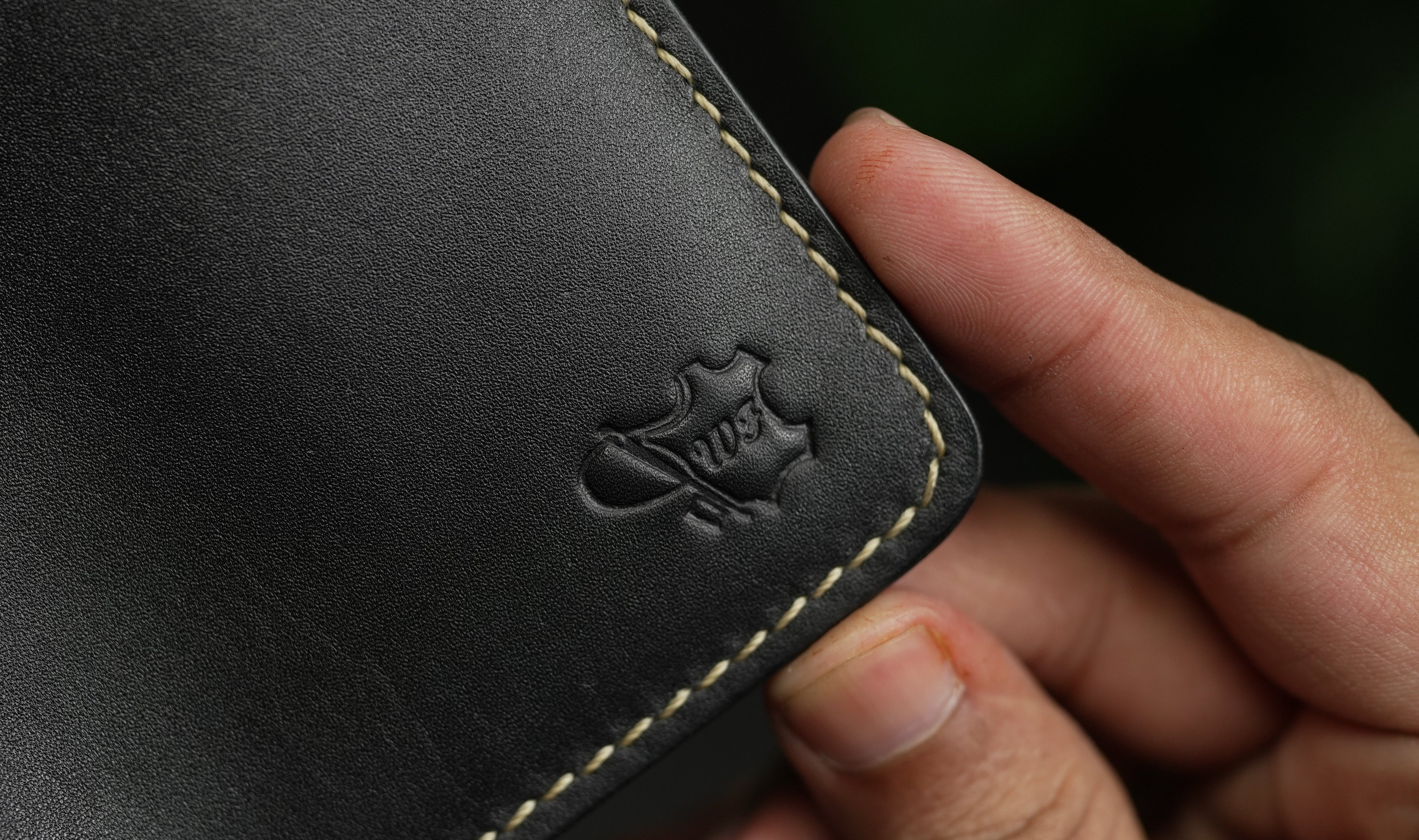 Close-up of a hand holding a black leather wallet with a brand logo on a dark background